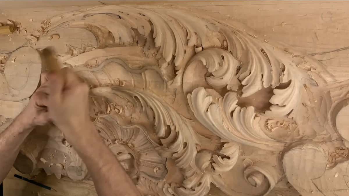 Both hands work a gouge at the left end of a long board of curling acanthus leaves.