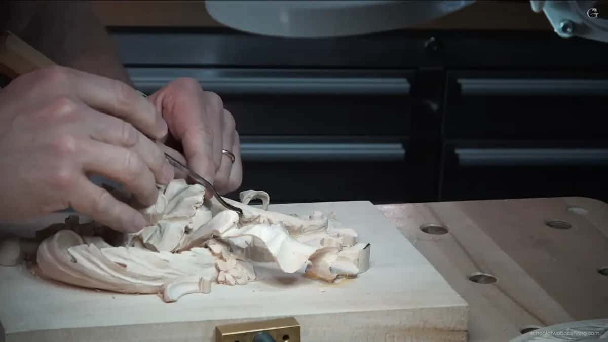 Side view of a bent tool going in under a leaf lobe, the carving lying flat on a board