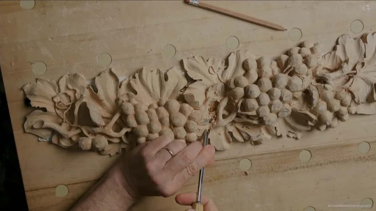 From overhead, one hand works a slim tool into the vine while a long pencil lies above the carving