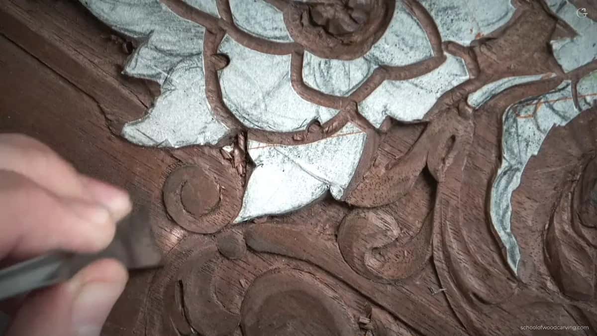 Fingers hold a gouge at the left side of the carved curl, chips around it
