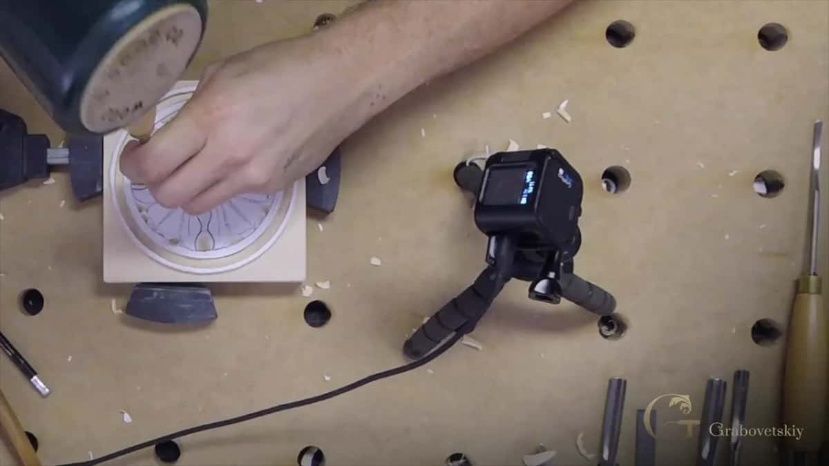 The mallet taps a gouge at the rim of a petal while a small camera on a tripod watches.