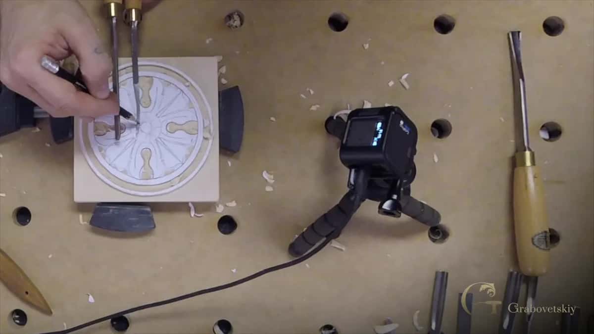 Over the drawing, one hand holds two tools and a pencil while the other steadies the block.
