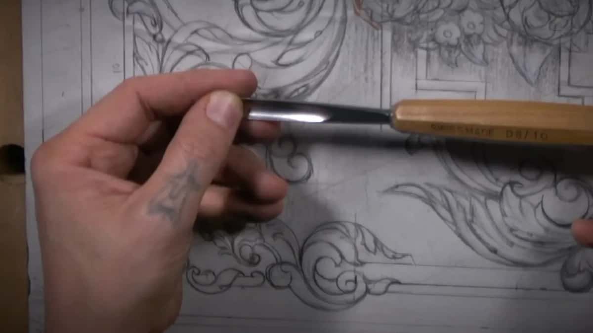 A long shiny gouge blade laid to a drawn curve, the tattoo on his wrist just showing