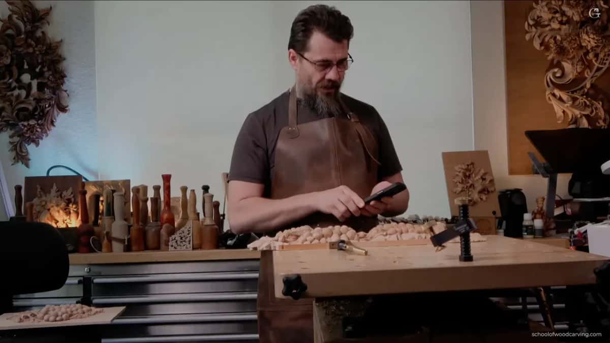 Standing at the bench in a leather apron, looking at a phone above the panel
