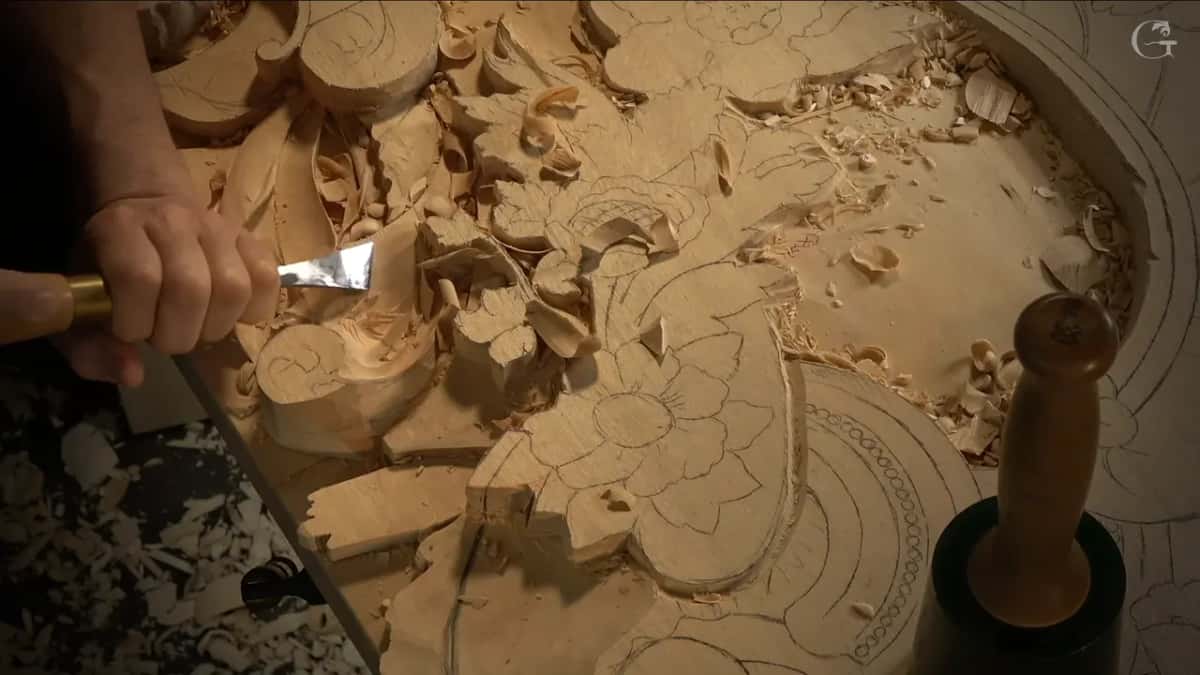 From above, one hand pushes a flat chisel along the curve, and a mallet stands upright at the right