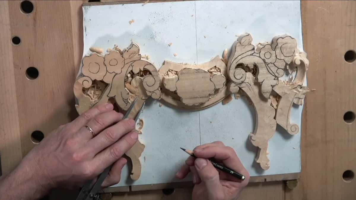 Calipers in one hand and a pencil in the other, held over the half-carved blank with its carved scrolls around it