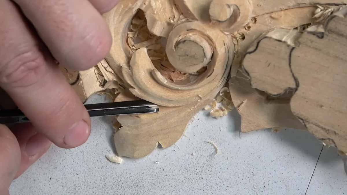 Slim gouge steered along the leaf below the scroll while chips lie scattered on the white board
