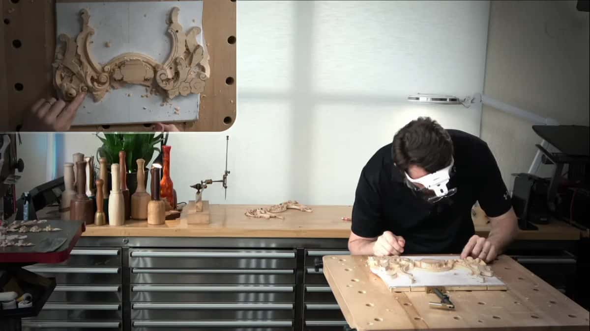 Leaning far over the panel at the bench, with a white magnifier on his head and both hands down at the work
