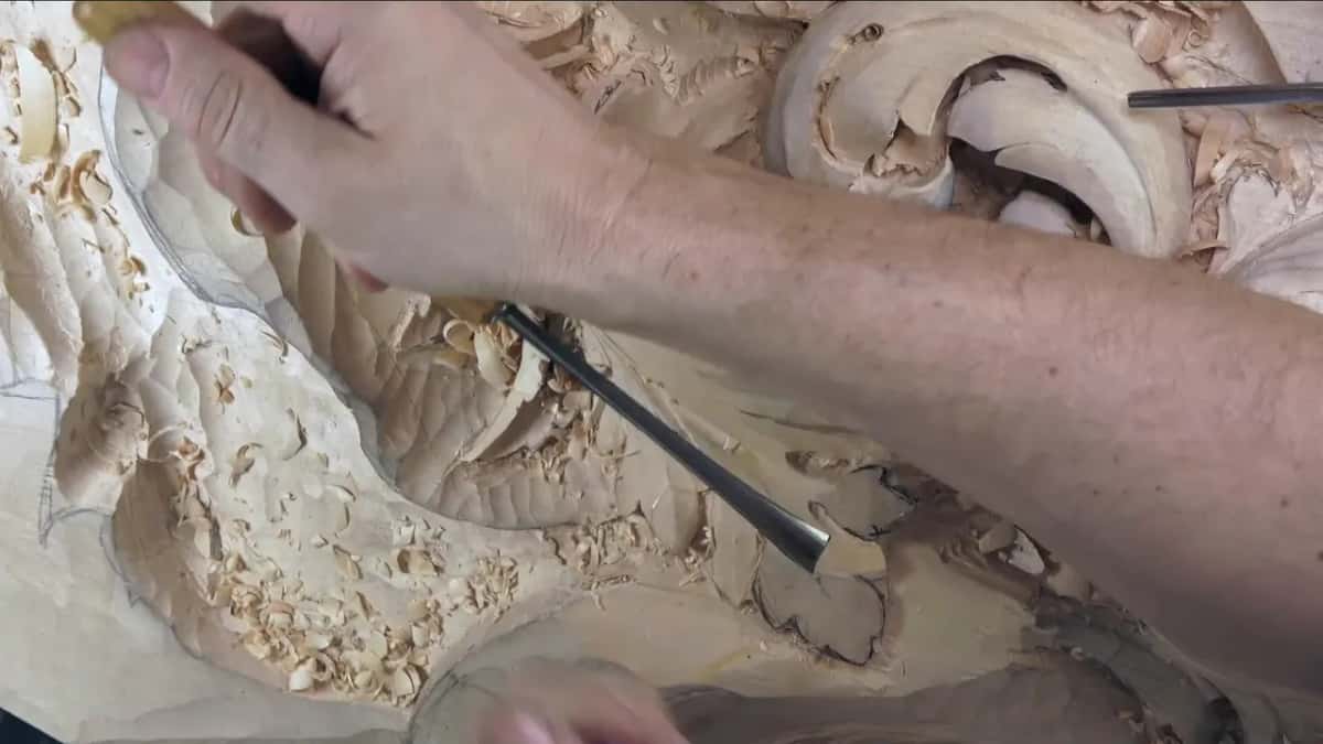 A bare forearm crosses the board as a gouge digs into the lower lobe of the leaf.