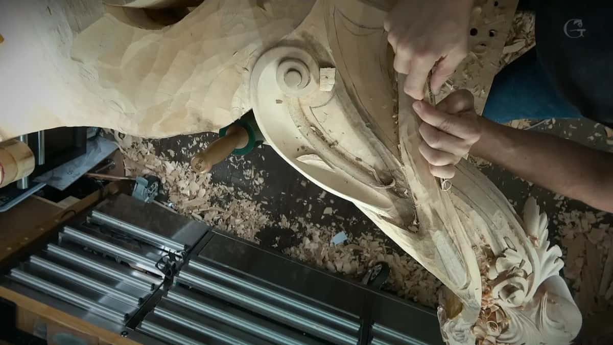Two hands grip a gouge high on the twisting band, the scroll to the left and bench rails below