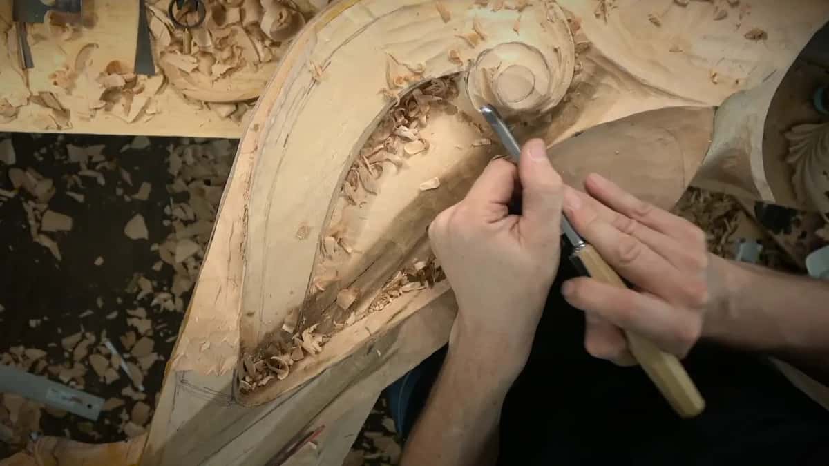 Right hand drives a gouge into the hollow beside the round volute while chips pile up in the groove