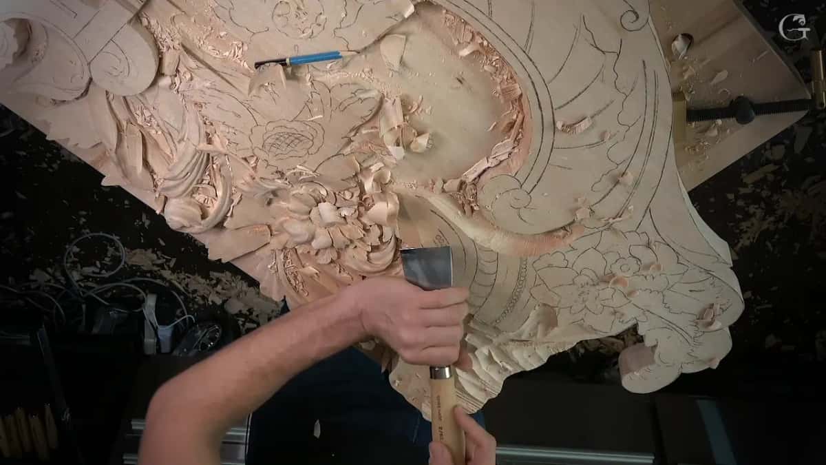 From above, two hands hold a broad gouge at the lower edge of the panel