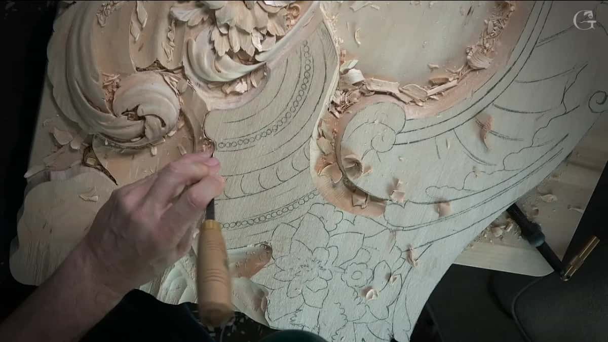 Seen from above, a thin tool rests in one hand beside the carved scroll and loose chips
