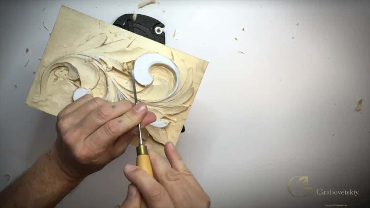 Thin tool held in both hands by the bottom curl of the tilted panel, chips around it