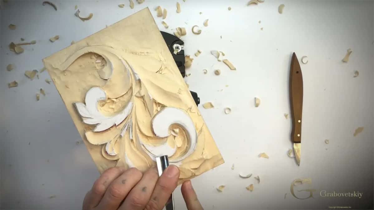 Gouge held low near the bottom of the leaf, wood chips scattered around and a knife lying to the right