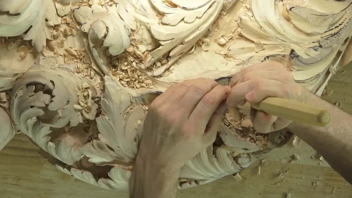 Both hands hold a gouge low over the scalloped edge of the carved board, chips all over it.