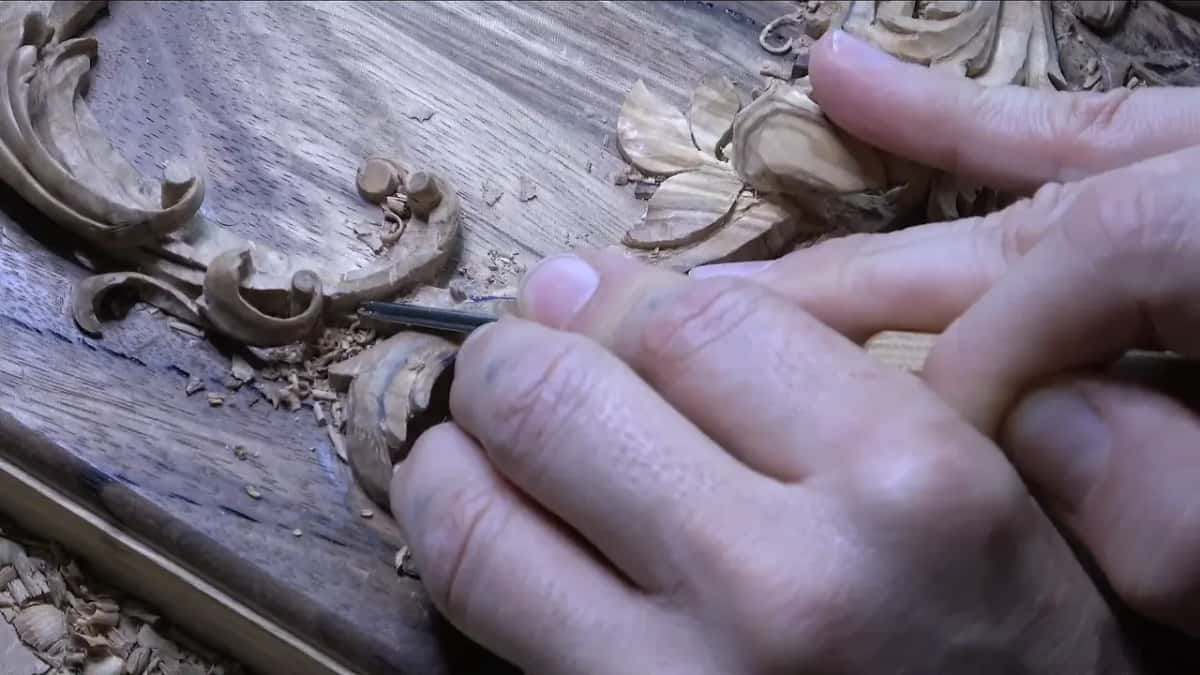 A thin tool slides under the edge of an olive leaf next to a volute, while the other hand steadies the blade