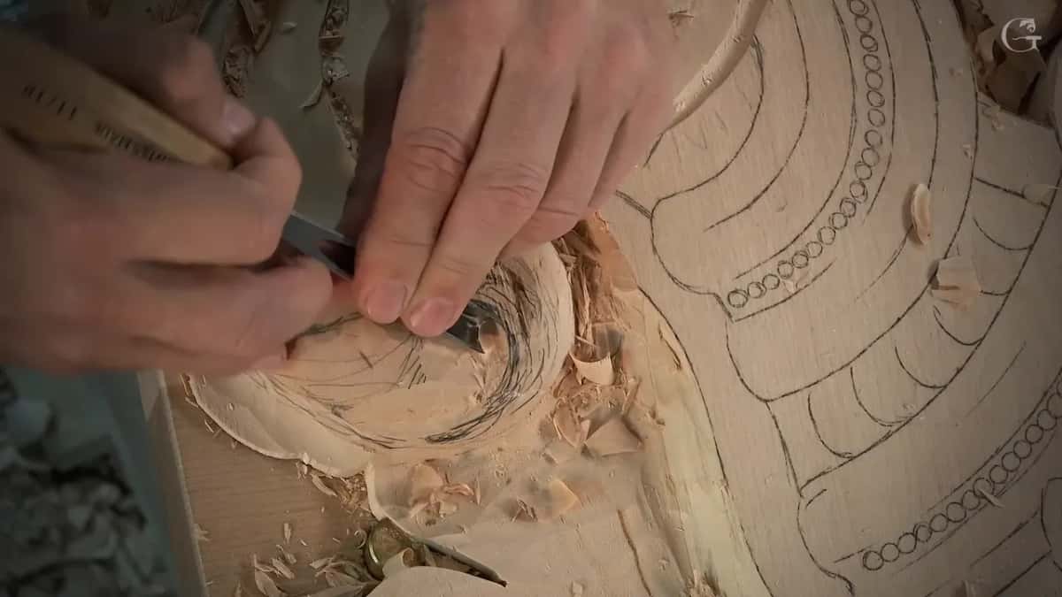 From another side, both hands press a small gouge into the pencil lines while shavings curl around it