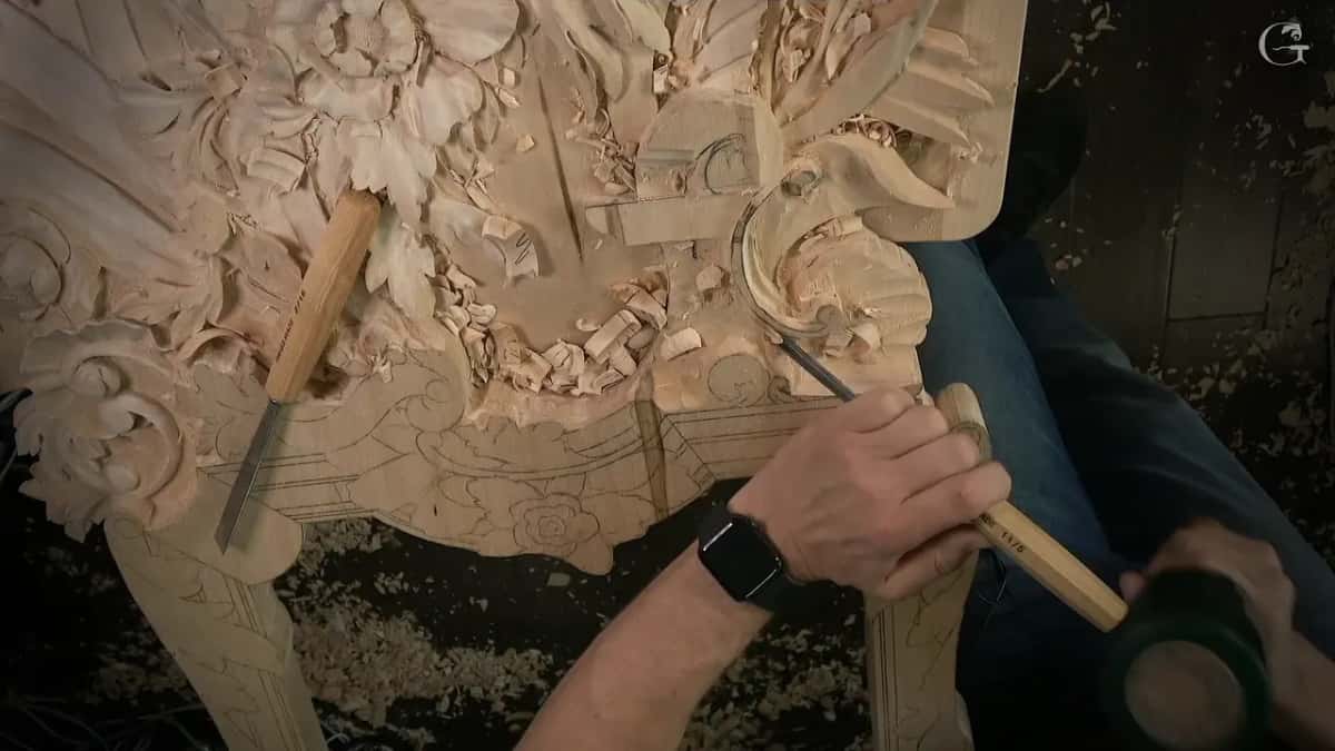 A wide flat tool rests on the carving while a slim gouge and a mallet work at the volute