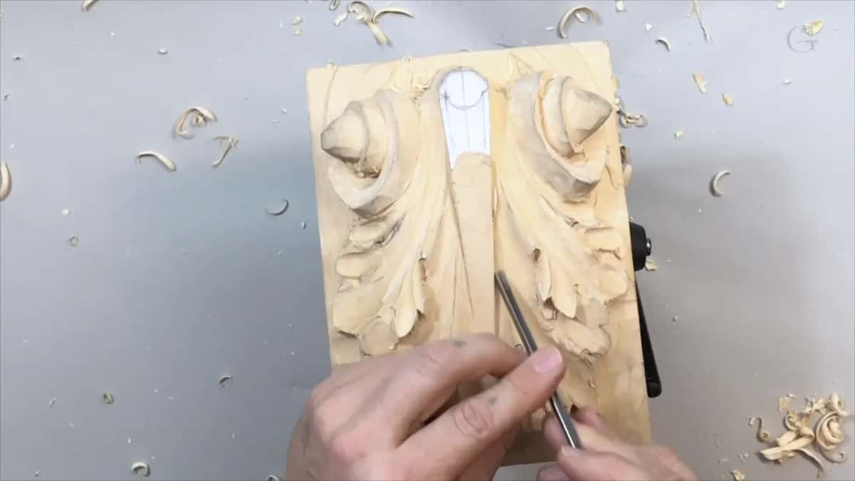 Both hands on a slim tool low on the right half of the leaf, chips curled on the bench
