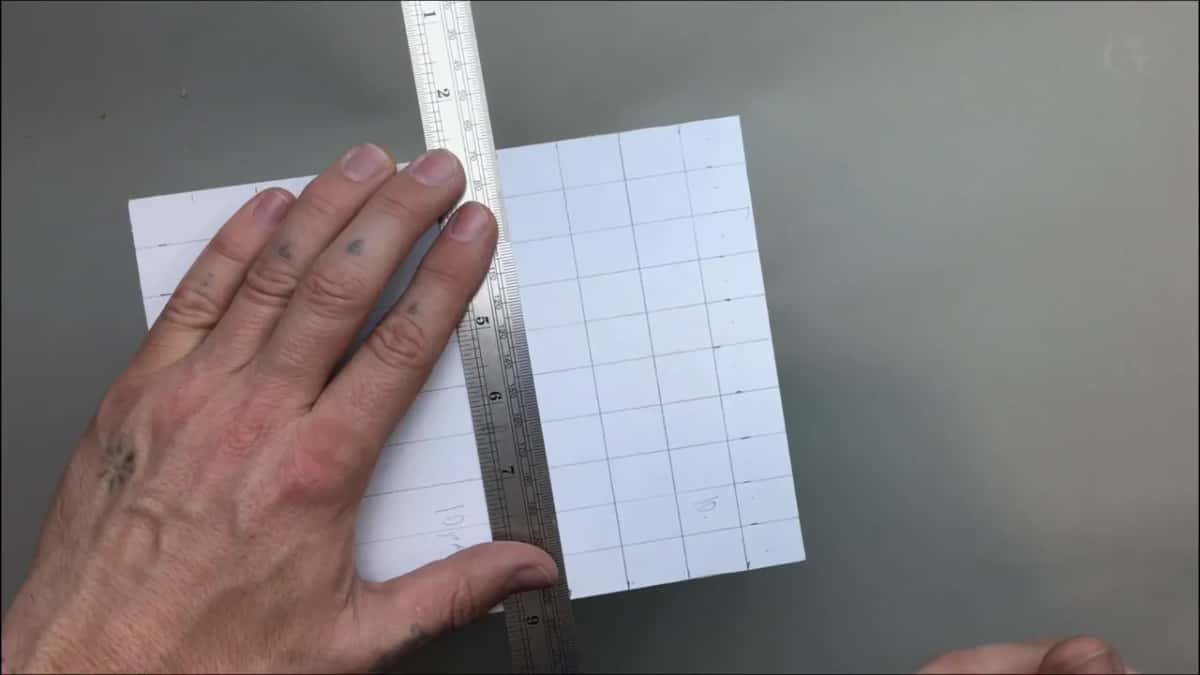 One hand presses an upright ruler flat on the grid, fingertips of the other hand at the bottom edge