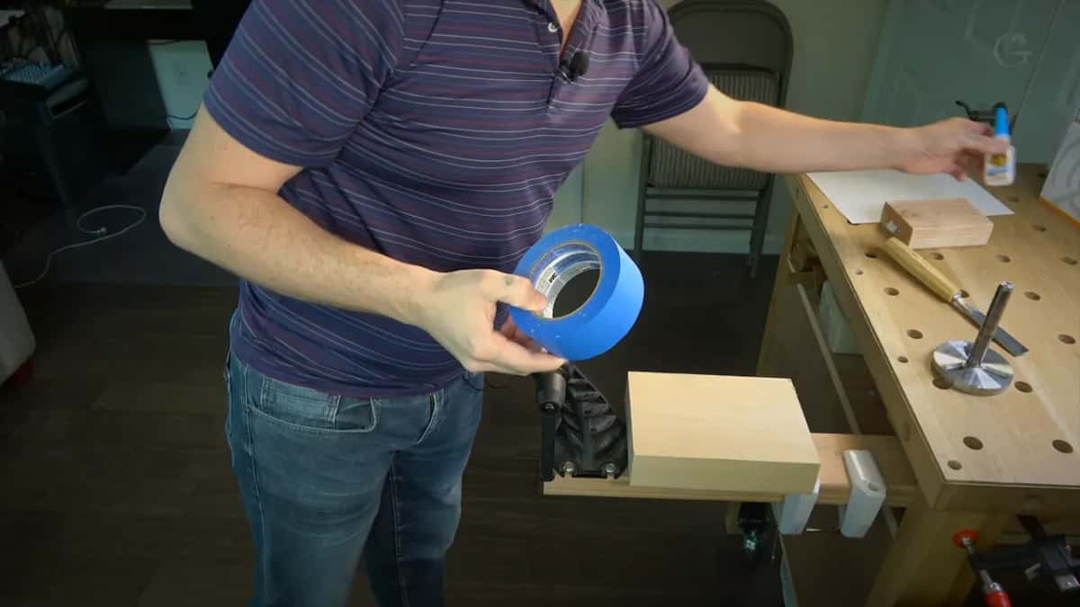 Roll of blue tape in one hand, the blank on the vise below, a small bottle on the bench