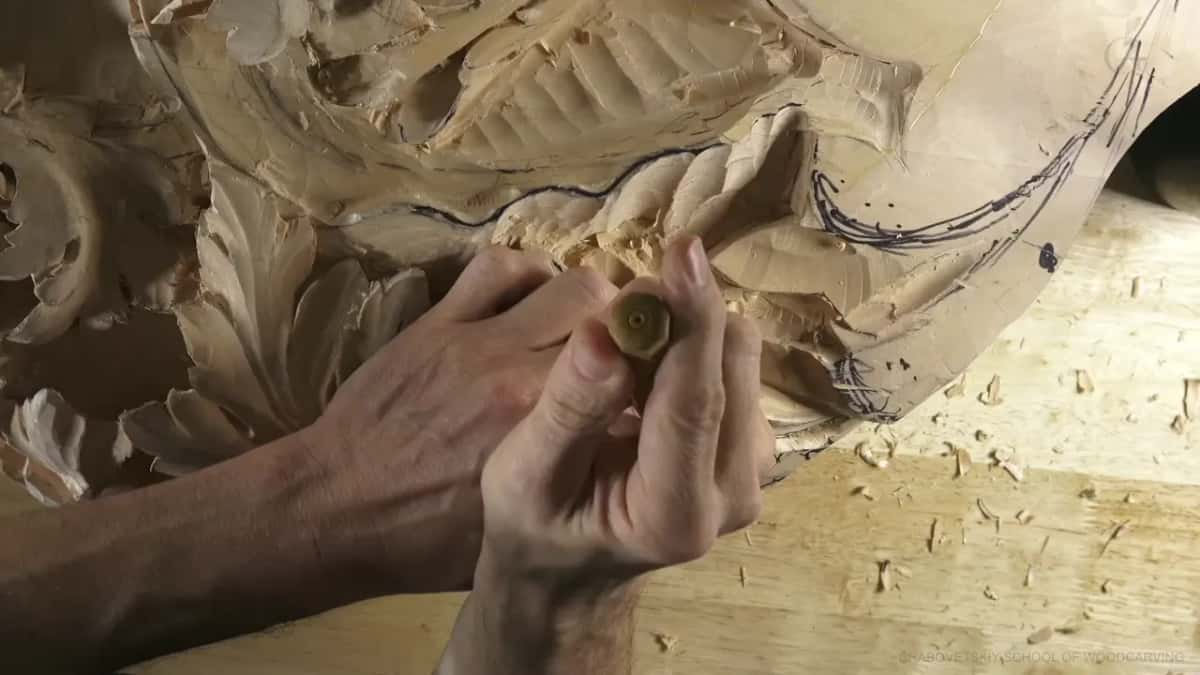 Two hands close over a short tool with a round handle end, its tip deep among the carved leaves.