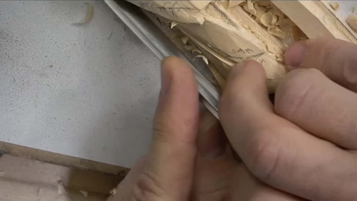Fingertips pinch a narrow tool beside the long molding, with shavings piled up in the leaf behind it.