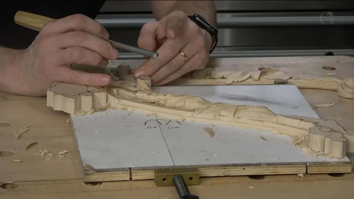 Both hands work the long carving on its white board, clamped at the front edge of the bench.