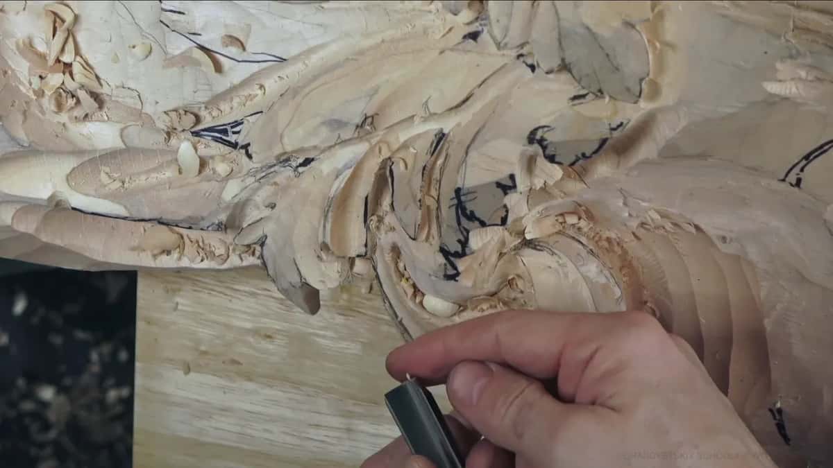 Pinched between two fingers, a short dark-handled tool works low at the base of the curl, near the bench.