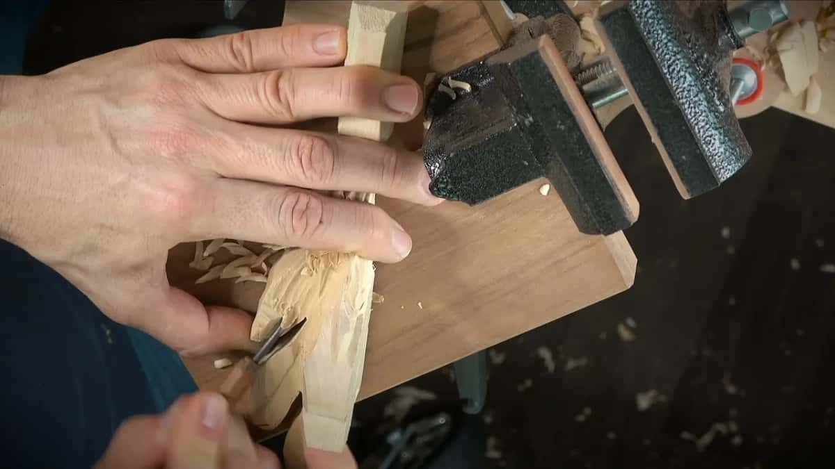 Fingers pin the block to the board as a small gouge scoops, shavings piling up beside the vise
