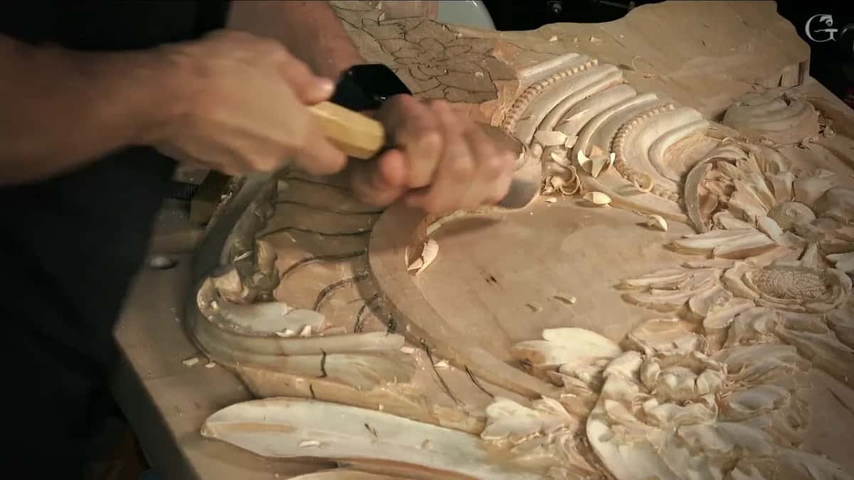 Seen low from the side, both hands push a gouge into the edge of the scroll