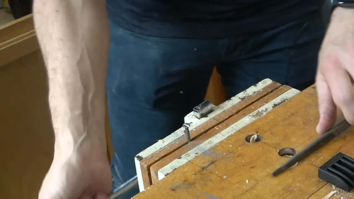 Short nail standing in a bench vise, a file resting on the wood beside it