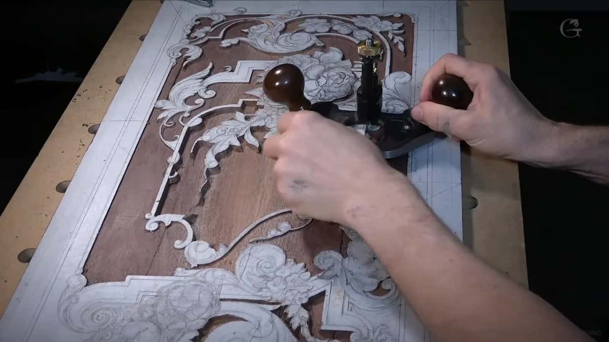 Both hands on the router plane over the board, the ornament still under white paper against brown wood