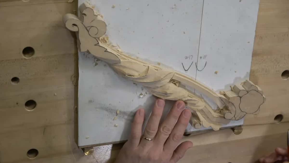 A hand rests flat on the white board beside the long acanthus carving, its flower end on the right.