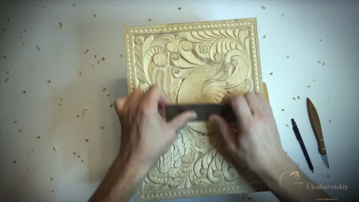 Scraper held in both hands across the panel, with a pencil and the knife lying on the bench
