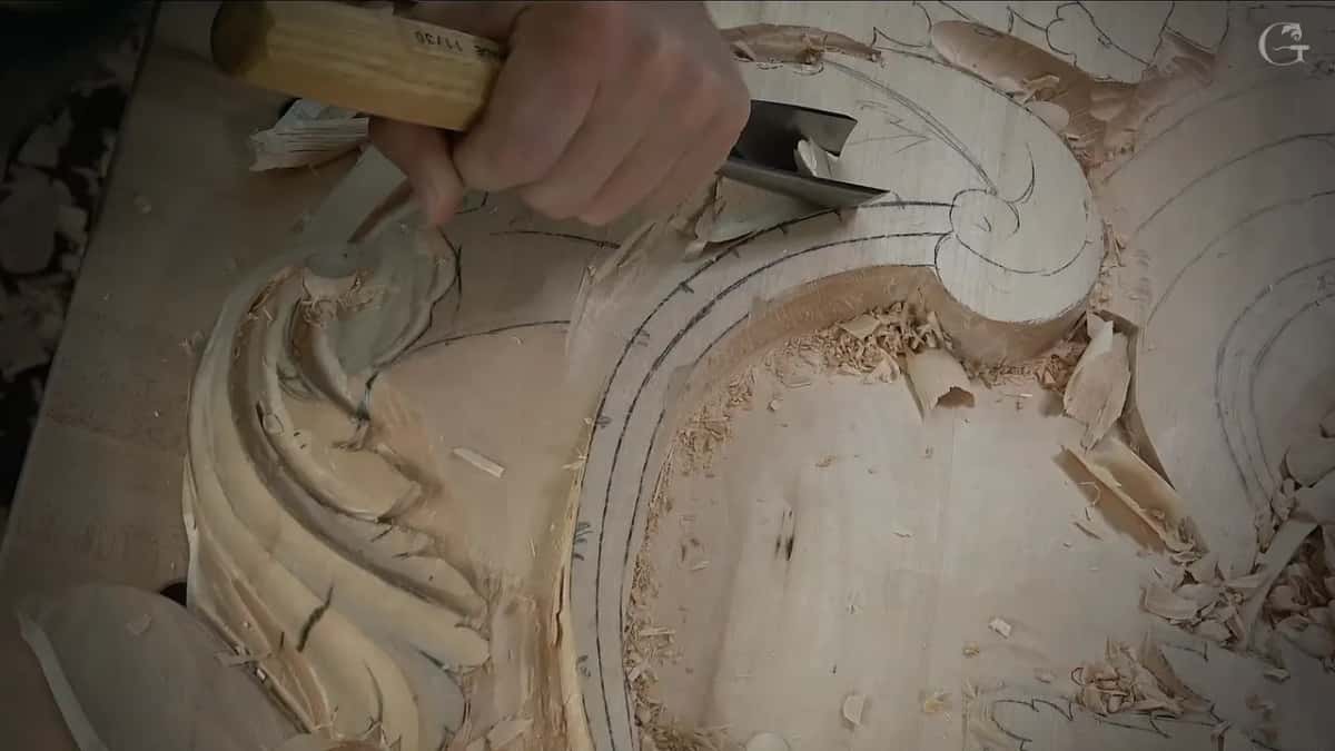 Right hand pushing a gouge along the drawn line beside a carved curl, shavings piled below