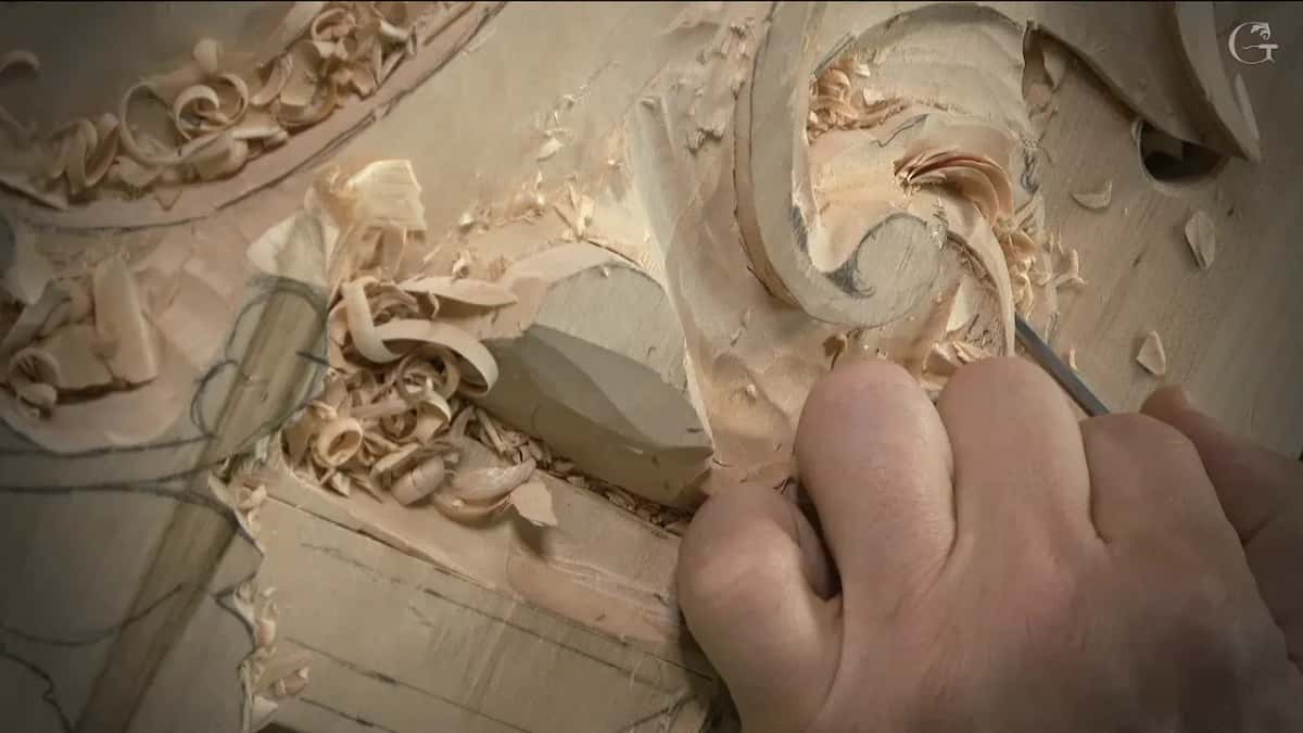 Under a tall scroll, a thin tool works beside straight pencil lines, shavings piled to the left