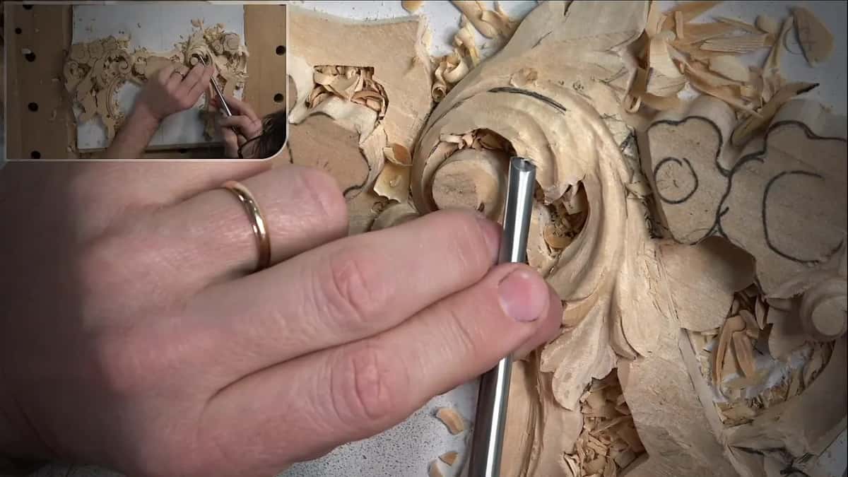 Steel gouge pressed into the leaf right of the scroll, its blade bright against the wood