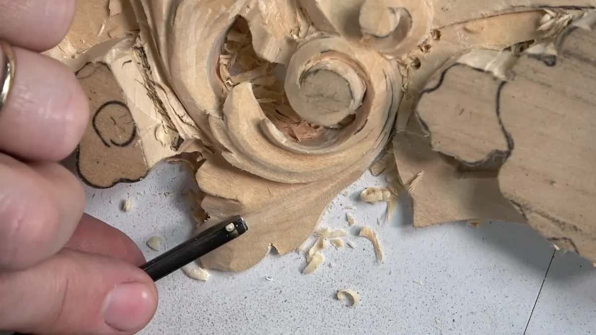 Black-handled tool held low at the leaf, under the curl of the scroll, with chips beside it