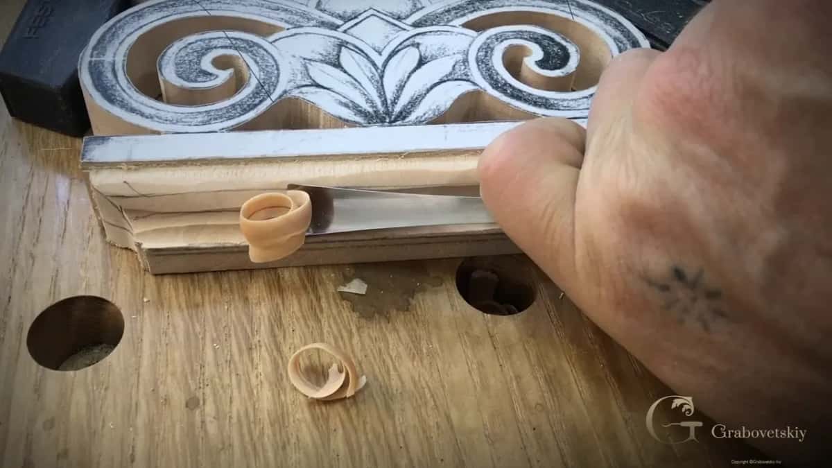 A gouge rides the stepped base of the acroterion and lifts one pale curl of basswood off the edge