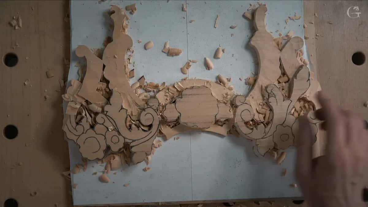 From overhead, half-roughed scrolls on white paper, wood chips scattered and a hand near the right edge