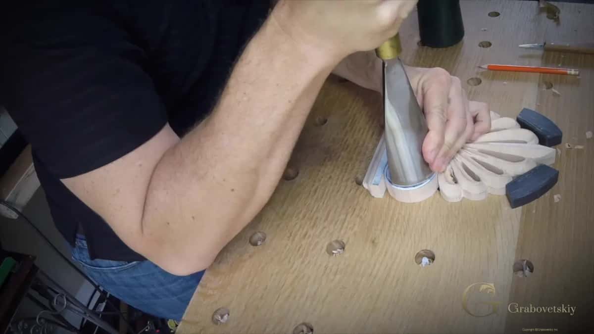 From the side, a wide gouge stands upright on the drawn spiral, one hand on the board