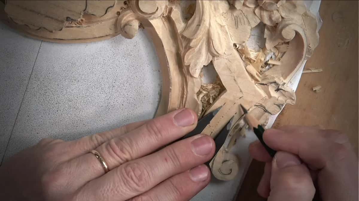 Black caliper jaws held across the molding in one hand, a sharp pencil marking it with the other