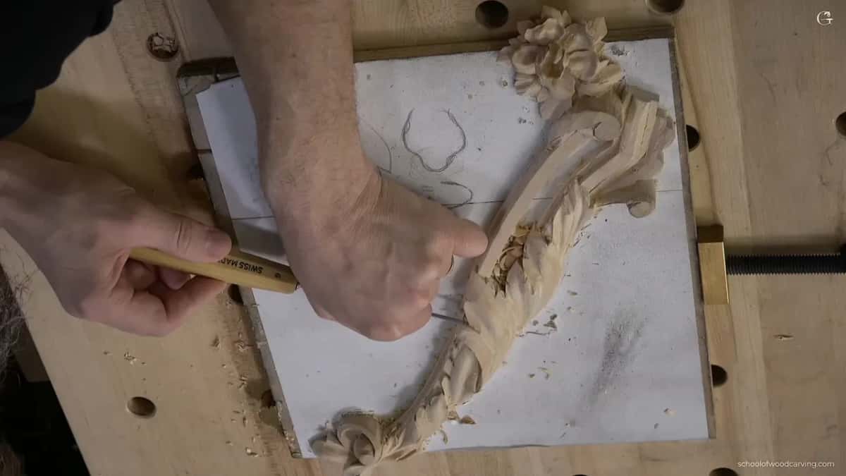 Two hands carve a long acanthus over a molding on a paper pattern, clamped to a bench with holes