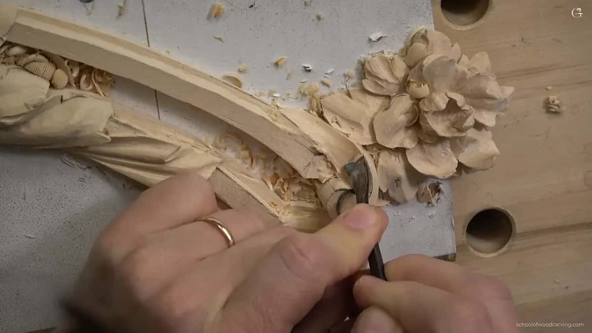 Close in, a gouge works between carved flower petals beside a curved molding, with chips all round