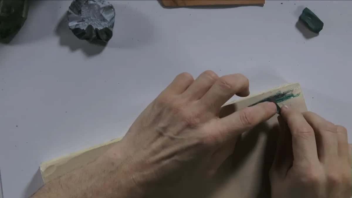 Green compound streaks a light leather strip, one hand holding it flat while the other hand presses down
