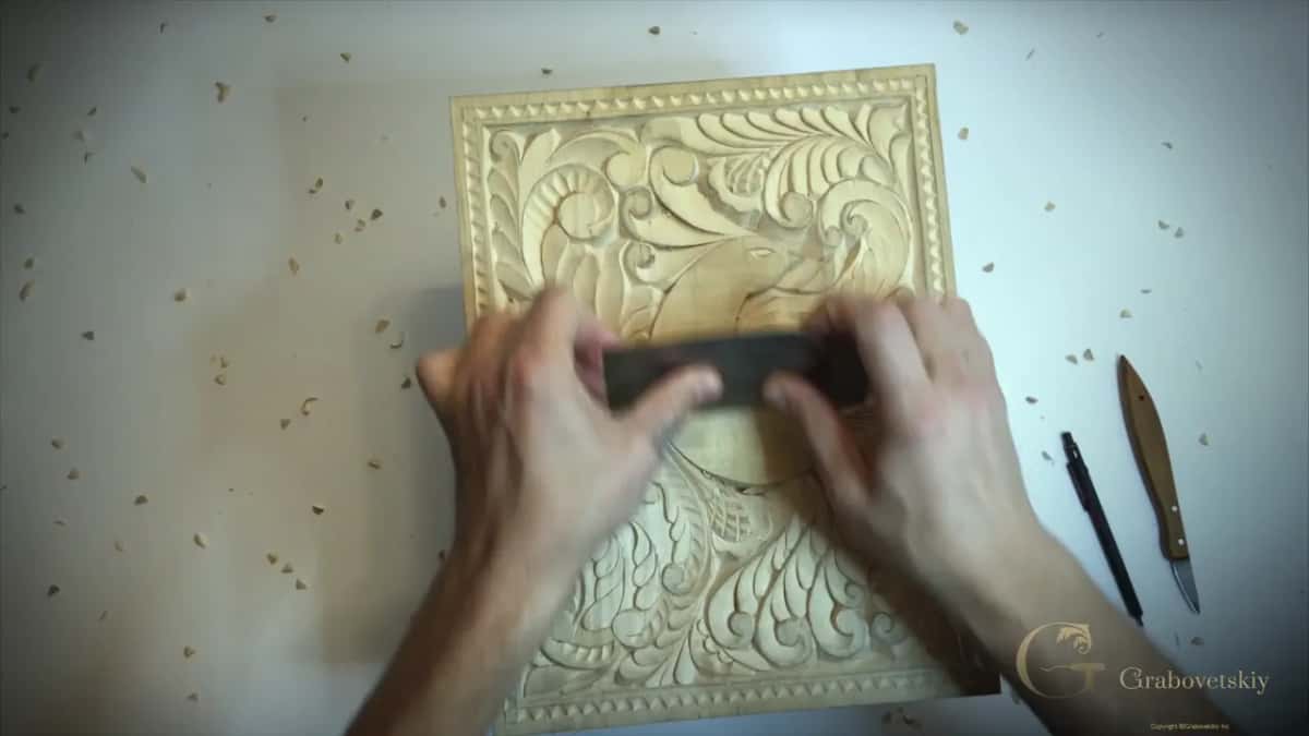 Both hands pull a steel scraper across the carved surface, a knife and pencil lying beside the board