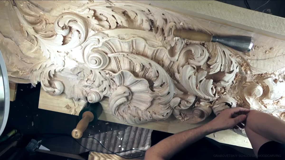 Long relief of scrolls and leaves lying flat, a gouge resting on top and two hands near the edge