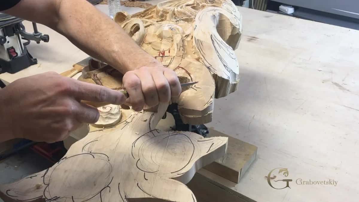 Both hands on one gouge at the edge of the big scroll, the left index finger pointing at the spot being carved